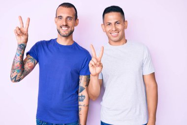 Gündelik giysiler giymiş, kameraya gülümseyen ve zafer işareti yapan iki genç gay. İki numara.. 