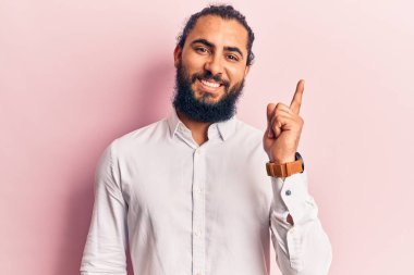 Yüzünde kocaman bir gülümseme olan sıradan kıyafetler giyen genç bir Arap, kameraya el ve parmak ile bakıyor.. 