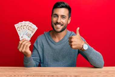 Yakışıklı İspanyol adam Yuan 'ın Çin banknotlarını tutuyor mutlu ve pozitif gülümsüyor, başparmağıyla mükemmel ve onay işareti yapıyor. 