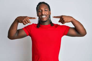 Saçları günlük kıyafetlerle örülü, neşeli gülümseyen ve parmaklarıyla ve ağzıyla işaret eden Afro-Amerikan bir adam. diş sağlığı kavramı. 