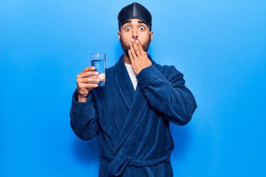 Uyku maskesi takan ve bornoz giyen genç İspanyol adam ağzını eliyle kapayarak su içiyor, şok geçiriyor ve hata yapmaktan korkuyor. şaşırmış ifade 
