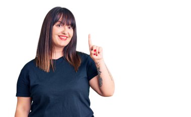 Genç ve cüsseli kadın gündelik kıyafetler giyer ve bir numaralı parmağını göstererek mutlu ve kendinden emin bir şekilde gülümser.. 