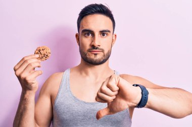 Sakallı, kolsuz tişörtlü, kızgın yüzlü, çikolatalı kurabiye yiyen genç yakışıklı bir adam. Negatif işaret baş parmakları aşağıda, reddedilme kavramı.