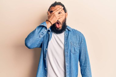 Gündelik giysiler giyen genç Arap adam şok içinde yüzünü ve gözlerini eliyle kapatıyor, parmaklarından korkuyla bakıyor. 