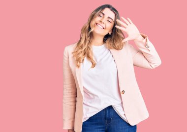 İş kıyafetleri giymiş genç beyaz bir kadın beş numarayı gösterip işaret ederken kendinden emin ve mutlu bir şekilde gülümsüyor.. 