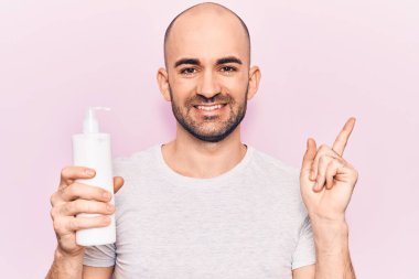 Genç yakışıklı kel adam elinde krem losyon şişesiyle gülümsüyor mutlu bir şekilde elini ve parmağını yana doğru gösteriyor. 