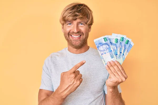 Joven Hombre Rubio Sosteniendo Billetes Rand Sudafricanos Sonriendo Feliz Señalando —  Fotos de Stock