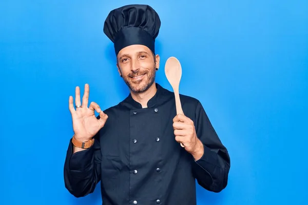 年轻英俊的男子身穿炊具制服 拿着勺子手拿着大勺 手拿着手签 微笑着表示友善的手势 是极好的象征 — 图库照片