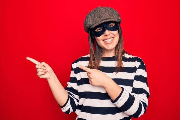年轻美丽的黑发窃贼女人 戴着帽子和面罩 站在孤立的红色背景上微笑着 看着摄像机 双手和手指指向旁边 — 图库照片