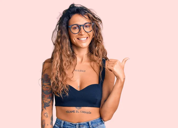 stock image Young hispanic woman with tattoo wearing casual clothes and glasses smiling with happy face looking and pointing to the side with thumb up. 