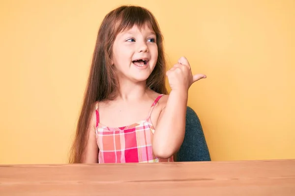 卡卡西亚小女孩 穿着休闲装 坐在桌子上 手指指向旁边 笑着张开了嘴 — 图库照片