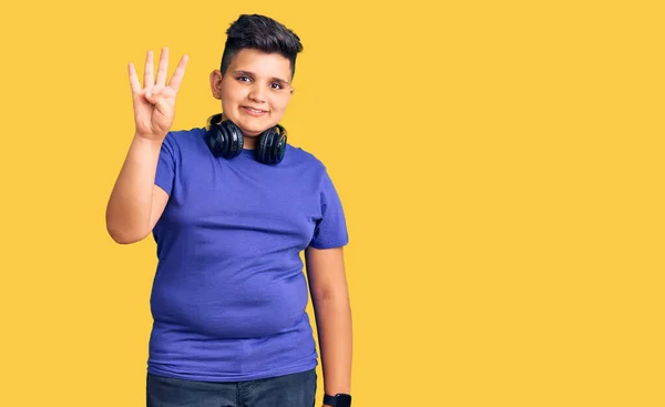 自信を持って幸せな笑顔ながら 小さな男の子は音楽を聞いてヘッドフォンを見せ 指の数4で指摘します — ストック写真