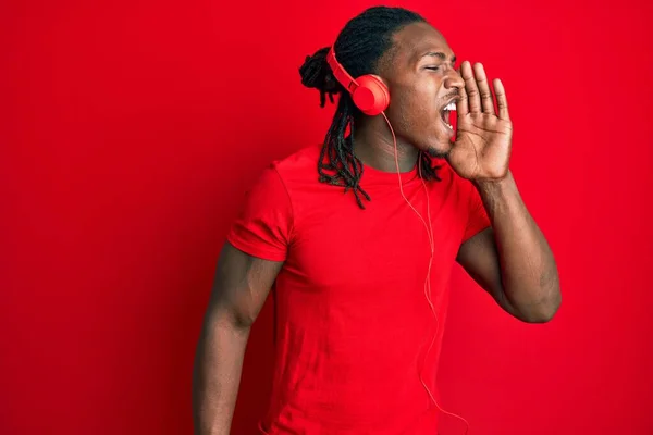 African american man with braids listening to music using headphones shouting and screaming loud to side with hand on mouth. communication concept.