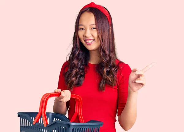 年轻美丽的中国姑娘牵着超市购物篮 笑容满面 手指手画脚地指向旁边 — 图库照片