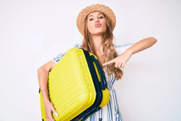 Young caucasian woman with blond hair wearing summer dress and holding cabin bag looking at the camera blowing a kiss being lovely and sexy. love expression.