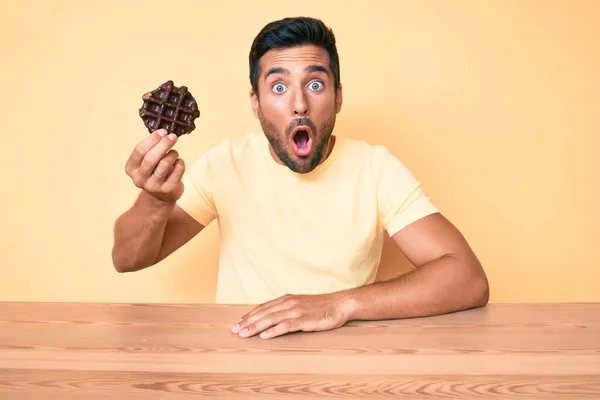 若いヒスパニック系の男チョコレートワッフルテーブルの上に座って怖がって驚きのために口を開けて驚いて 不信の顔 — ストック写真