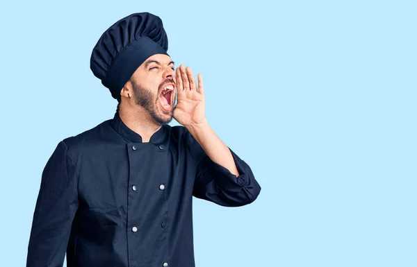Young hispanic man wearing cooker uniform shouting and screaming loud to side with hand on mouth. communication concept.