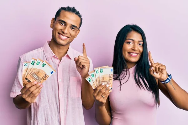 Young latin couple holding 50 euro banknotes smiling with an idea or question pointing finger with happy face, number one