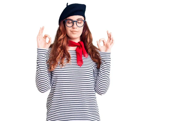 Fransız tarzı giyinen genç ve güzel bir kadın. Bere takmış, gevşemiş ve gözleri kapalı gülümsüyor. Parmaklarıyla meditasyon hareketi yapıyor. yoga konsepti. 