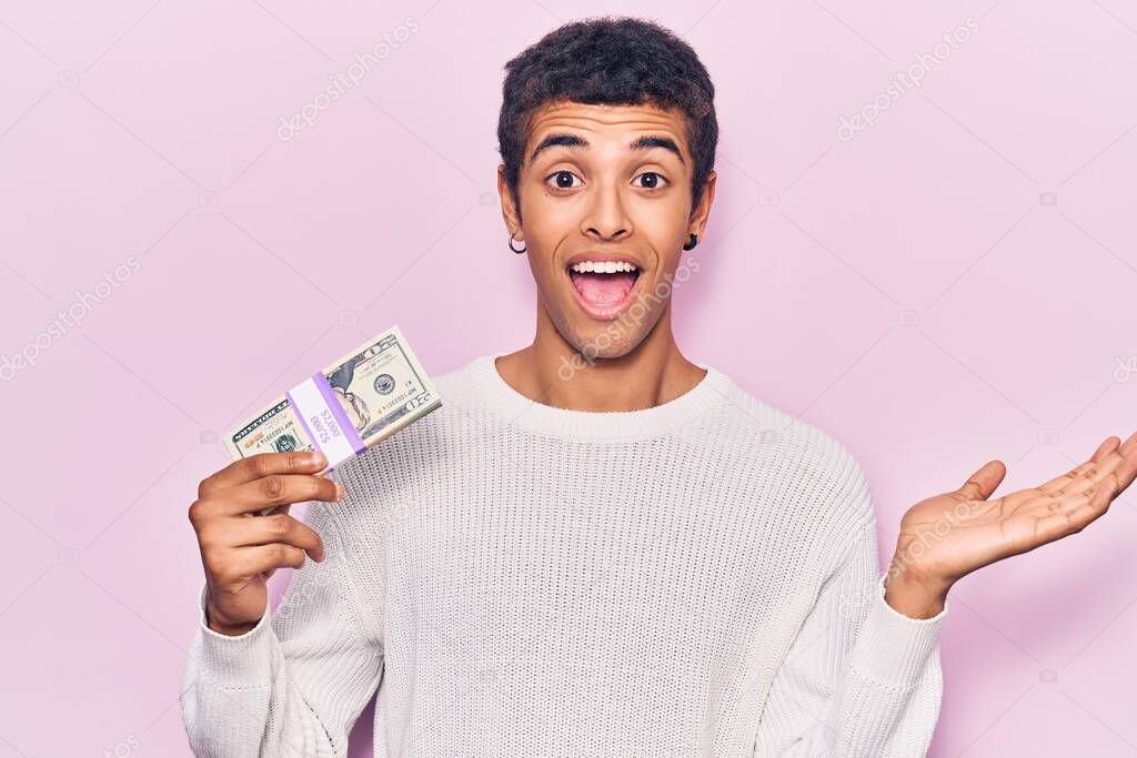 Young african amercian man holding dollars celebrating achievement with happy smile and winner expression with raised hand 