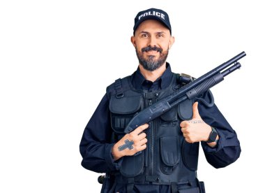Polis üniforması giyen genç ve yakışıklı adam elinde tüfekle mutlu ve pozitif gülümsüyor, başparmağıyla mükemmel ve onay işareti yapıyor. 