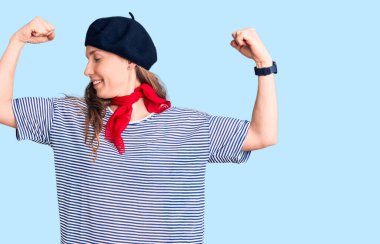 Fransız beresi ve çizgili tişört giyen genç, güzel sarışın kadın, gururla gülümseyen kol kaslarını gösteriyor. fitness konsepti. 