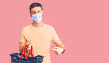 Alışveriş sepeti ve tıbbi maske takan yakışıklı genç adam mutlu bir şekilde elini ve parmağını gösterdi. 