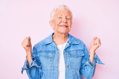 Mavi gözlü, gri saçlı, sıradan bir kot ceket giyen son sınıf güzeli bir kadın. Çok mutlu ve heyecanlı. Kolları havada, gülümseyen ve başarı için çığlık atan bir jest yapıyor. kutlama konsepti. 