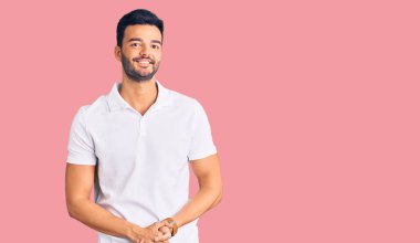 Genç, yakışıklı, İspanyol bir adam, günlük kıyafetler giyip ellerini birleştiriyor ve gülümsüyor. Rahatlamış ve neşeli. Başarı ve iyimserlik 