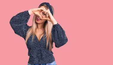 Genç, güzel, sarışın, gündelik kıyafetler giyen, kalp şeklinde el ve parmaklarıyla gülümseyen, tabeladan bakan bir kadın. 