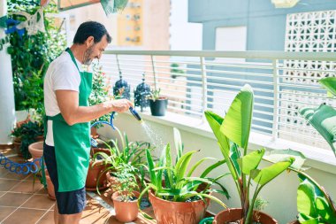 Orta yaşlı sakallı adam gülümsüyor mutlu bir şekilde terasta çiçekleri suluyor.