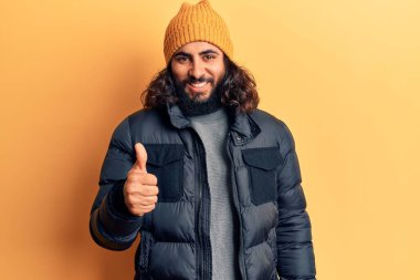 Gündelik kışlık kıyafetler giyen genç Arap adam mutlu ve pozitif gülümsüyor, başparmağıyla mükemmel ve onay işareti yapıyor. 