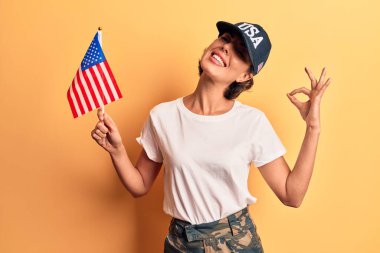 Amerika Birleşik Devletleri bayrağı taşıyan güzel bir kadın parmaklarıyla işaret veriyor, gülümseyerek mükemmel bir sembol oluşturuyor. 