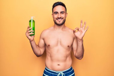 Tatilde sakallı, mayo giyen, elinde aloe vera kremi şişesi tutan yakışıklı bir adam.
