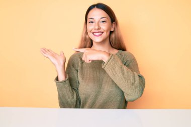 Genç esmer kadın, gündelik giysiler giyip masanın üzerinde şaşkın şaşkın oturuyor ve kameraya gülümsüyor.. 