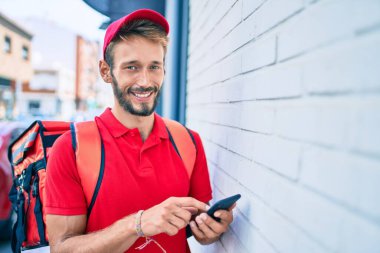 Beyaz teslimatçı kırmızı üniforma giyiyor ve sırt çantası teslim ederken gülümsüyor ve akıllı telefon kullanıyor.