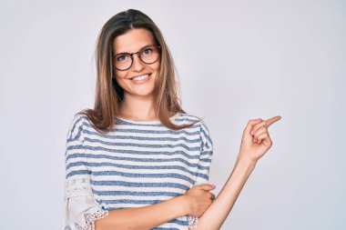 Güzel, beyaz bir kadın, günlük kıyafetler ve gözlük takıyor, gülümsüyor, mutlu bir şekilde elini ve parmağını yana uzatıyor. 