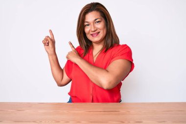 Orta yaşlı esmer kadın, gündelik kıyafetler giymiş masaya oturmuş gülümsüyor ve kameraya bakıyor. İki eliyle ve parmaklarıyla işaret ediyor.. 