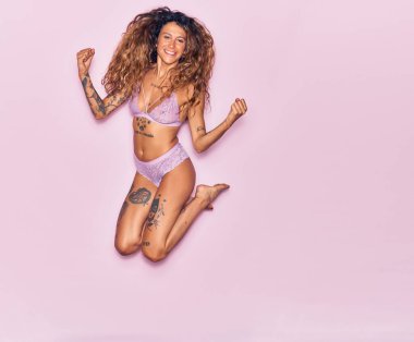 Dövmeli, iç çamaşırlı, mutlu gülümseyen genç bir kadın. Yüzünde tebessümle zıplıyor, pembe arka planda yumruklar havada kutluyor.