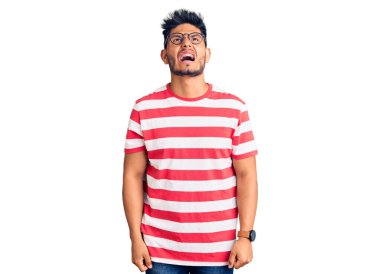 Yakışıklı Latin Amerikalı genç adam gündelik kıyafetler giyiyor ve gözlük takıyor kızgın ve kızgın bağırıyor sinirli ve öfkeli, öfkeyle bağırıyor. öfke ve agresif konsept. 