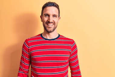 Sıradan çizgili tişört giyen genç yakışıklı adam mutlu ve havalı bir gülümsemeyle izole edilmiş sarı arka planda dikiliyor. Şanslı kişi..