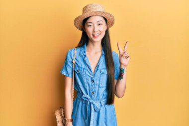 Genç Çinli kadın yaz şapkası takıyor ve iki numaralı parmaklarıyla işaret ederken kendinden emin ve mutlu bir şekilde gülümsüyor.. 