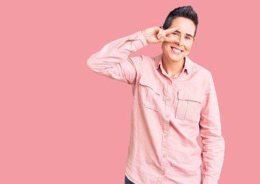 Kısa saçlı, günlük kıyafetler giyen, barış sembolü yapan, yüzünde parmakları olan, gülümseyen, zaferi gösteren genç bir kadın. 