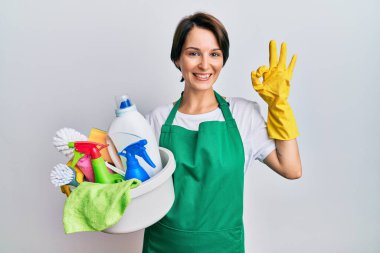 Kısa saçlı esmer kadın önlük giyip temizlik malzemelerini tutarken gülümsüyor ve el ve parmaklarıyla 