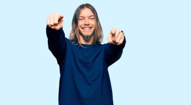 Uzun saçlı yakışıklı beyaz adam, günlük kışlık süveter giyiyor. Seni ve kamerayı parmaklarıyla gösteriyor, neşeli ve pozitif gülümsüyor. 