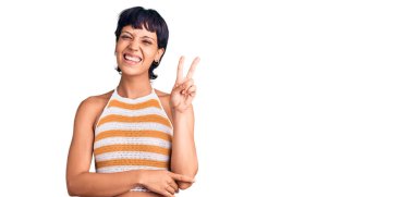 Kısa saçlı, sıradan kıyafetler giyen, gülümseyen, kameraya göz kırpan, parmaklarıyla zafer işareti yapan esmer bir kadın. İki numara.. 