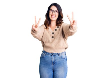 Güzel, genç, esmer bir kadın, günlük kıyafetler ve gözlük takıyor, gülümsüyor ve iki elini de zafer işareti yaparken gösteriyor. İki numara.. 