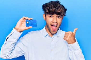 Genç İspanyol bir adam kredi kartı tutuyor. Parmağını yana doğru gösteriyor. Ağzını açıp gülümsüyor. 