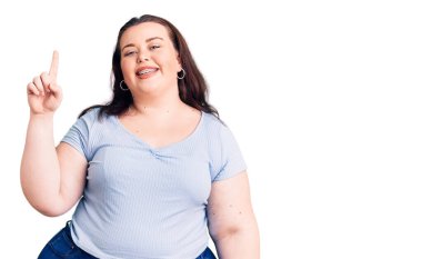 Genç ve cüsseli kadın gündelik kıyafetler giyer ve bir numaralı parmağını göstererek mutlu ve kendinden emin bir şekilde gülümser.. 