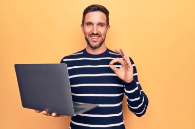 Genç yakışıklı adam dizüstü bilgisayar kullanarak izole edilmiş sarı arka planda duruyor tamam işareti yapıyor parmaklarıyla, gülümseyerek mükemmel bir sembol oluşturuyor.